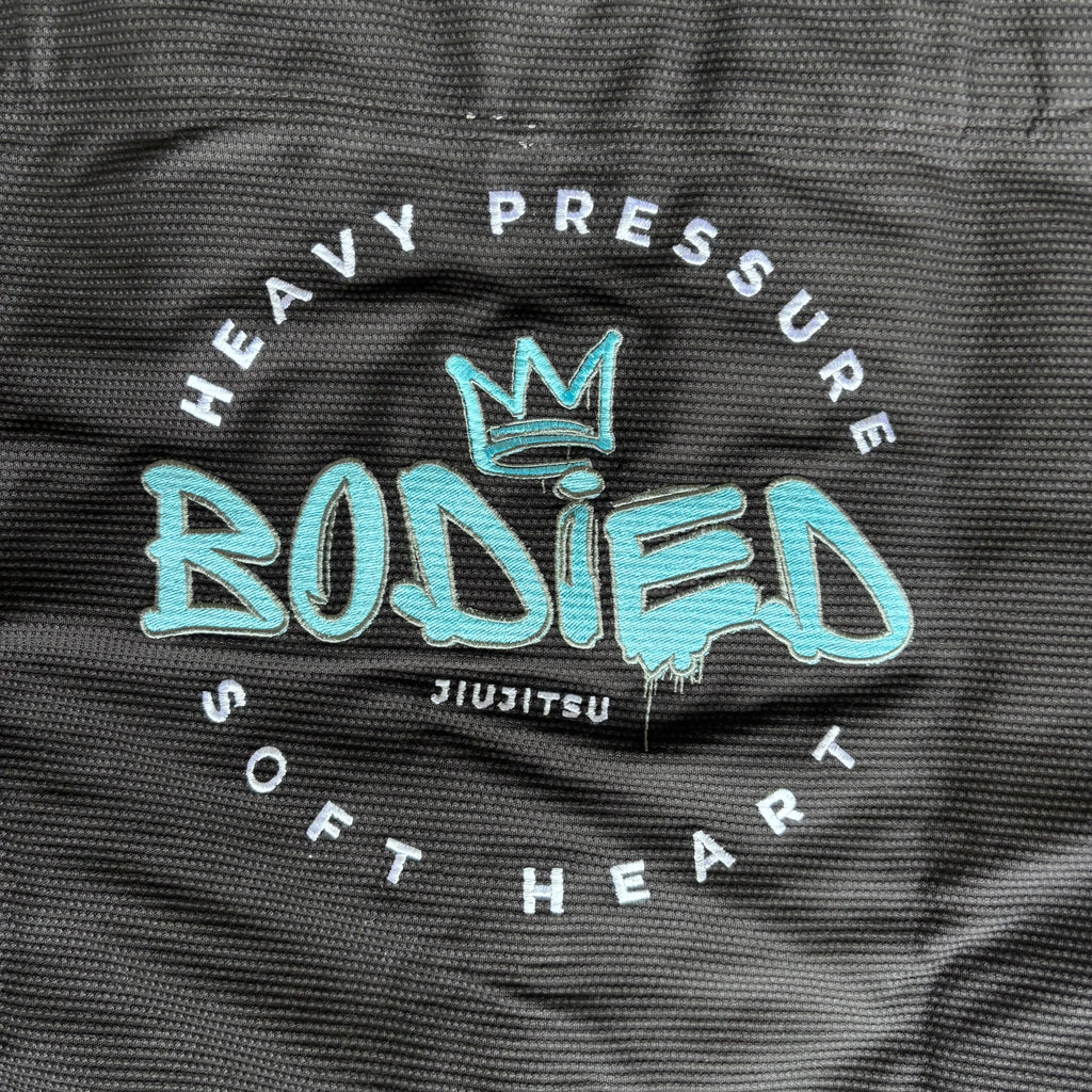 Close-up of a black textured fabric with a printed graphic reading 'HEAVY PRESSURE BODIED JIUJITSU SOFT HEART' in white and turquoise.